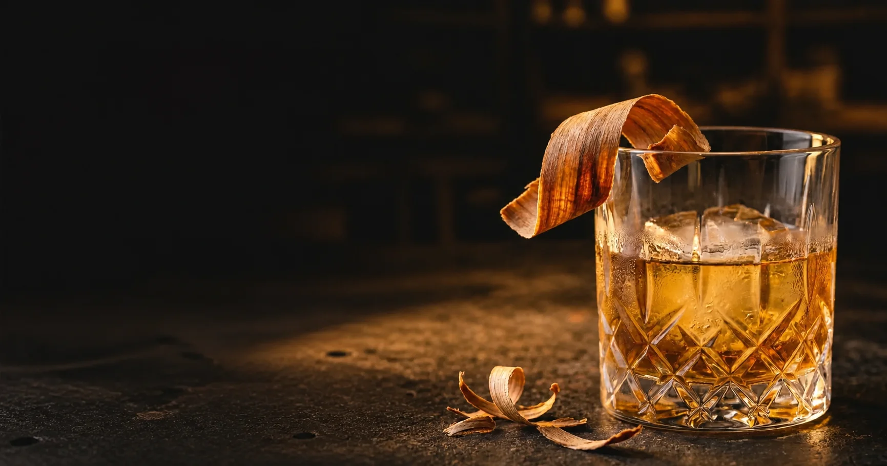 A whiskey glass with a curled cedar shaving on a dark workshop bench